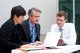 3 business people sitting at a table, looking at a laptop [Image by creator pkchai from AdobeStock]